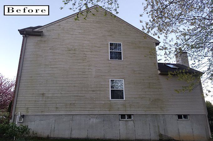 Before photo of a tan-sided house with a gray foundation and two windows under trees