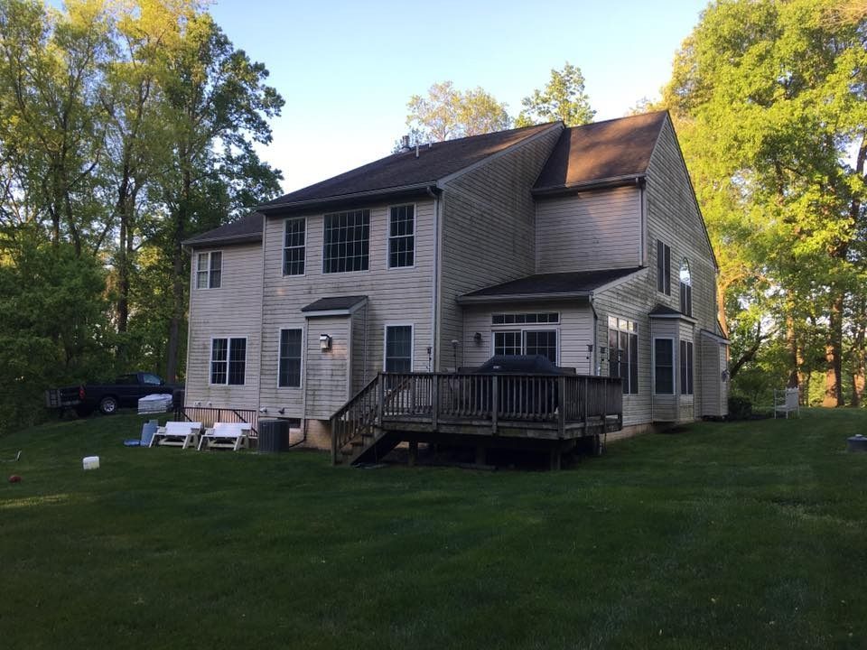 Two-story beige house with a dark deck and green lawn, surrounded by trees
