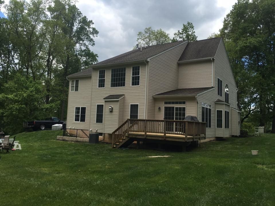 Two-story beige house with dark roof and wooden deck in a grassy yard, surrounded by trees