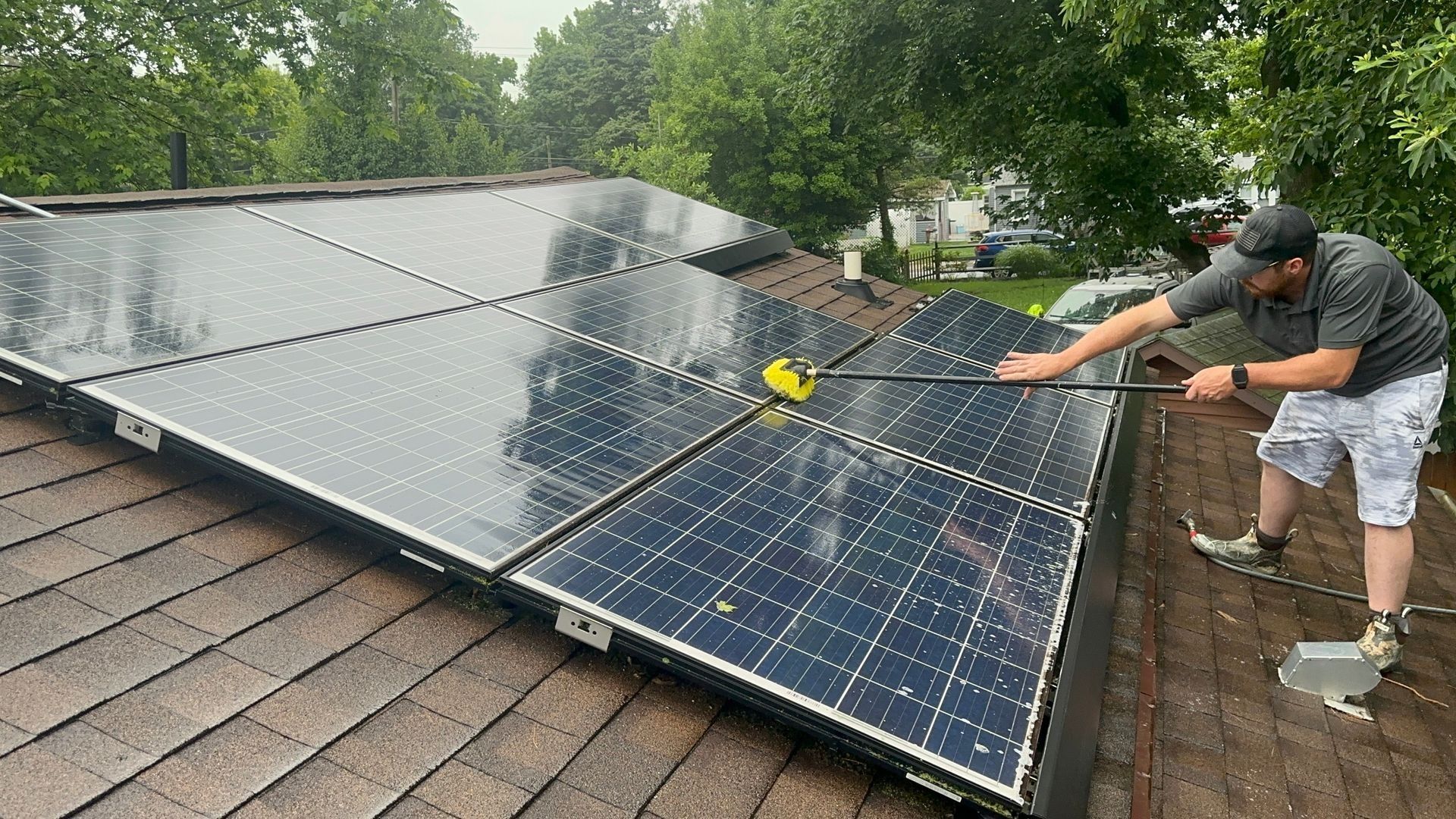 Workers cleaning solar panels on a shingled roof with a yellow brush, surrounded by trees.
