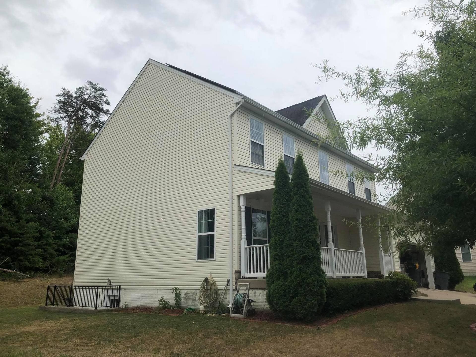 Two-story beige house with white trim and a small front porch, surrounded by trees and greenery