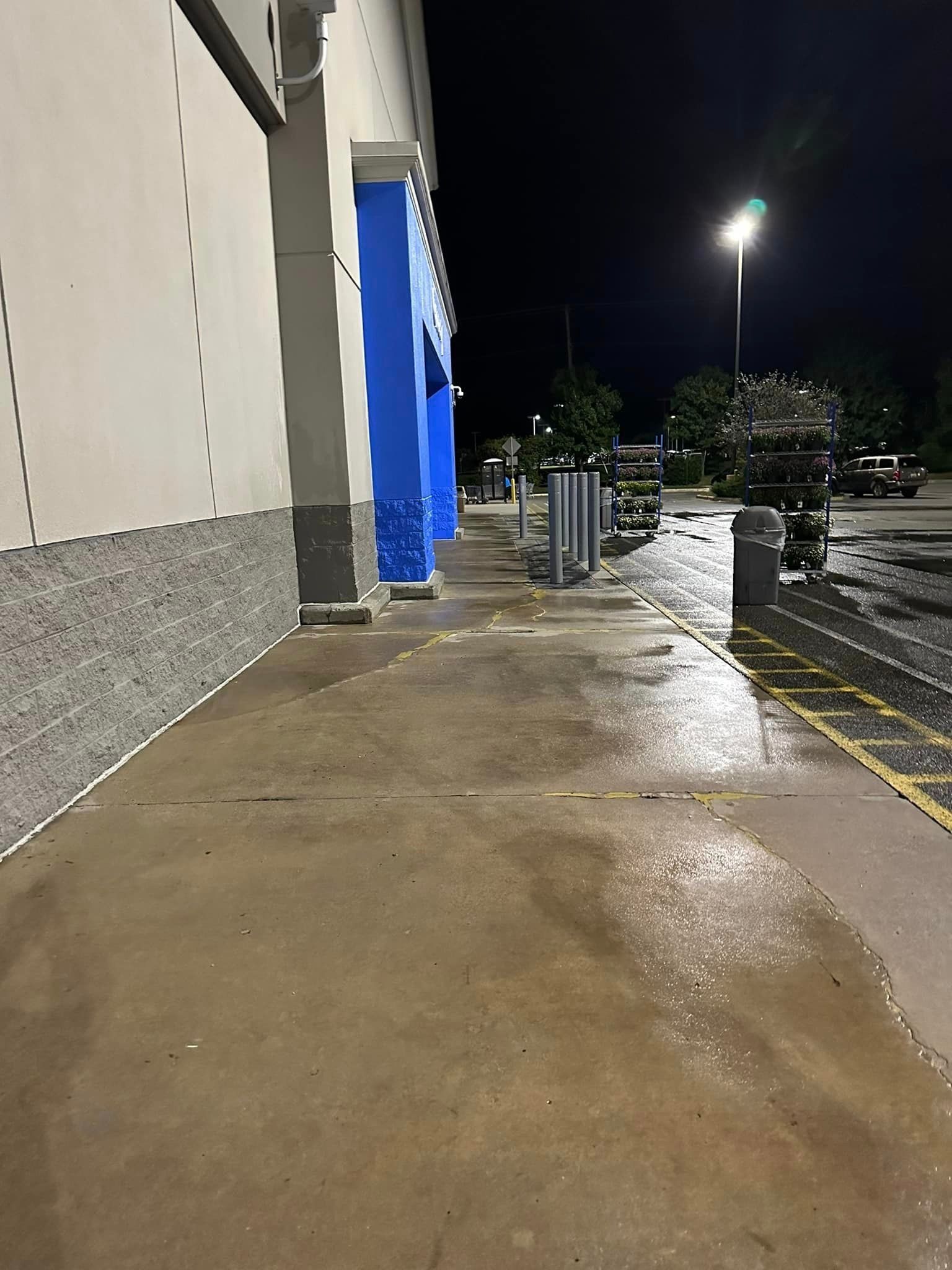 Wet sidewalk beside a building at night, with blue tarp-covered carts and parking lot lights nearby