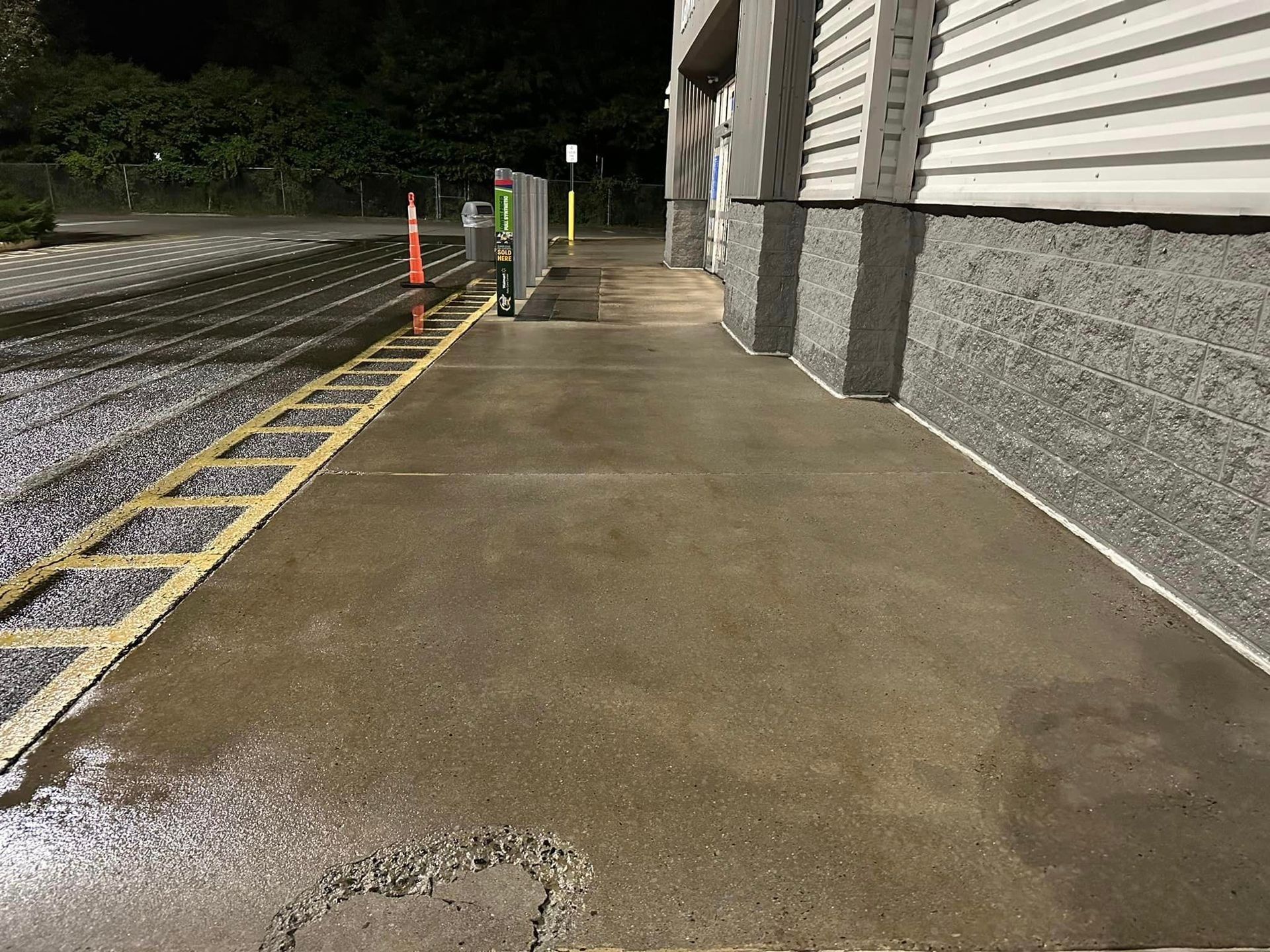Wet concrete sidewalk beside a building at night, with parking lot markings and orange cones ahead