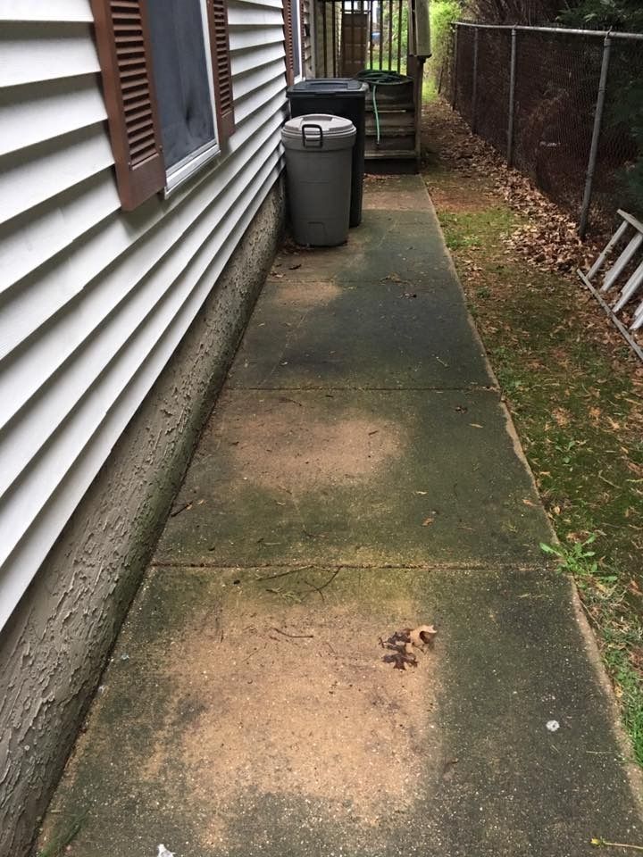 Narrow concrete side yard beside a house, with trash bins and a fence along the walkway.