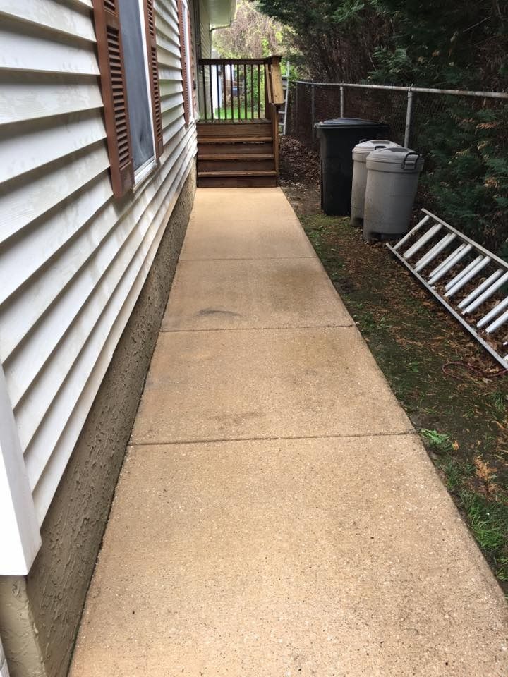 Narrow concrete walkway beside a house, with stairs, trash bins, and a ladder along the right side.