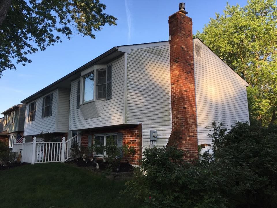 Two-story white house with a brick chimney, front porch, and shrubs in a sunny yard