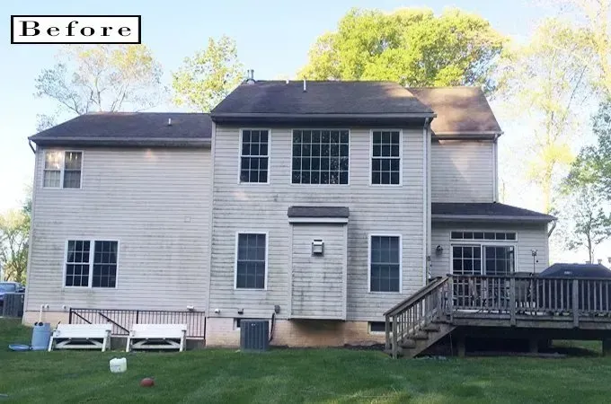 Before view of a two-story white house with a dark roof, front porch, deck, and backyard lawn