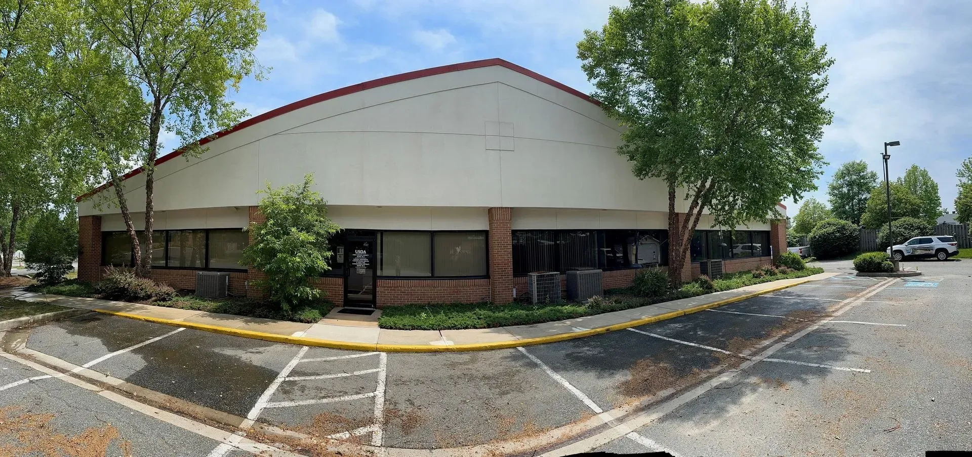 Low, tan commercial building with brick base, trees, and a mostly empty parking lot on a sunny day