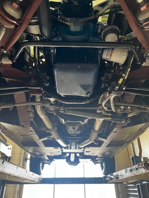 Underside of a classic car on a lift, showing engine, exhaust, and chassis details.