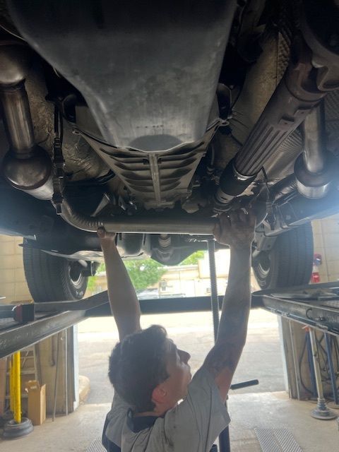 Mechanic working under a vehicle on a lift, reaching to adjust exhaust pipes, at a repair shop.