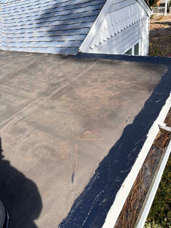 Flat roof with dark gray surface, edged with black sealant, next to a gutter filled with debris.