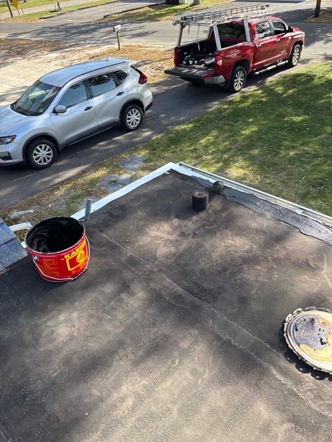 A flat roof with a bucket, vent pipe, and parked silver SUV and red truck.