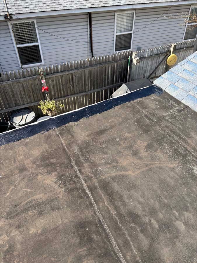 Flat, weathered roof with dark trim, overlooking a backyard with a fence and a gray building.