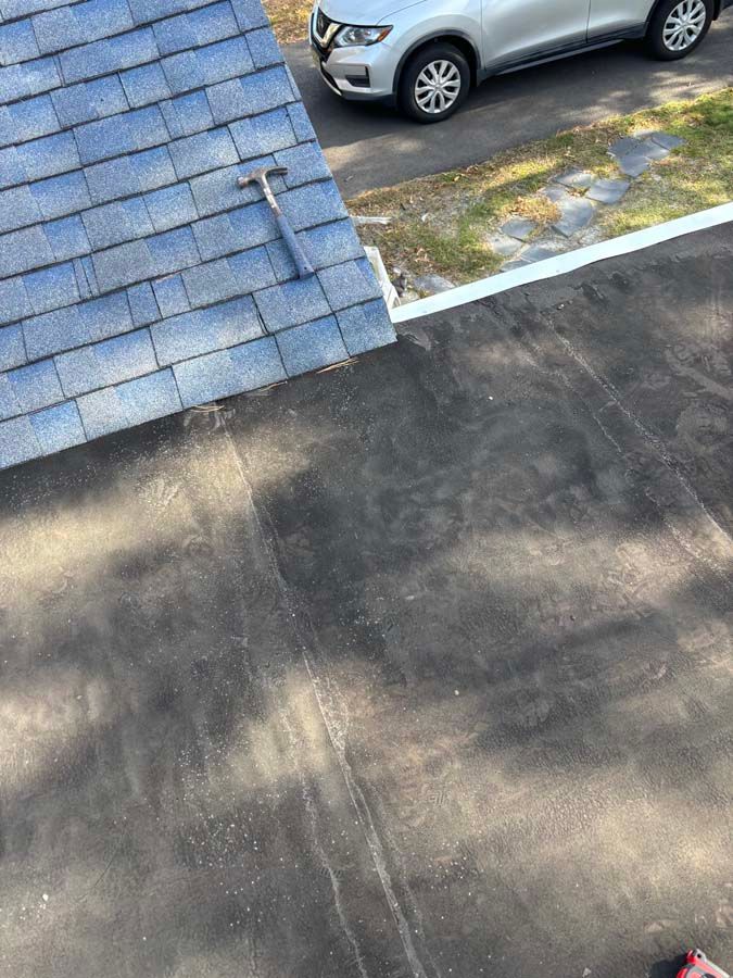 View of a dark, flat roof meeting a blue shingle roof. A silver car is parked nearby. A hammer rests on the shingles.