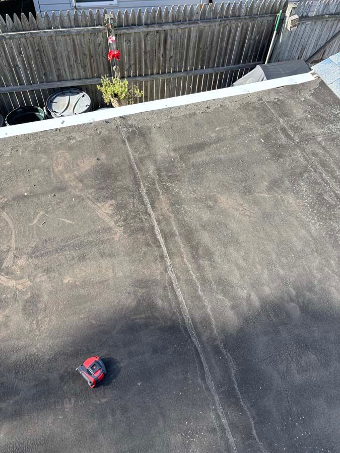Overhead view of a flat black roof with visible seams, a fence, and a red tool.