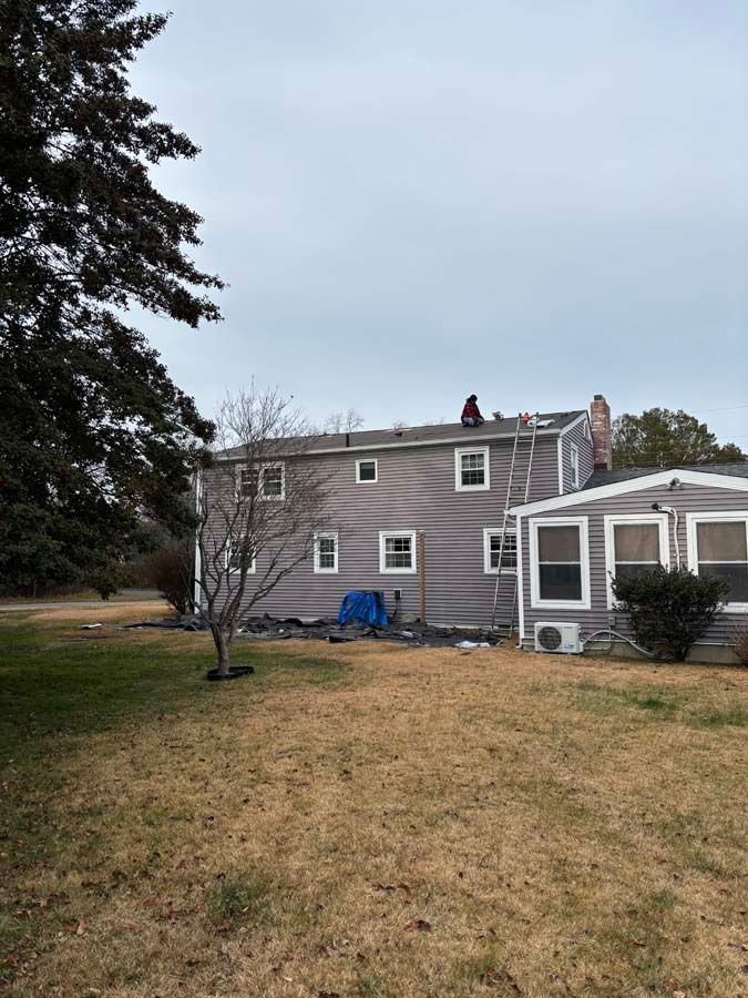Person on a roof repairing shingles. Gray house, ladder, cloudy sky, green grass.