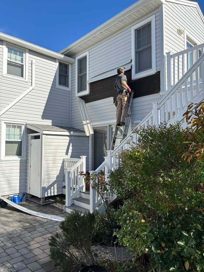 Man on ladder installing siding on a two-story white house with stairs and a small shed.