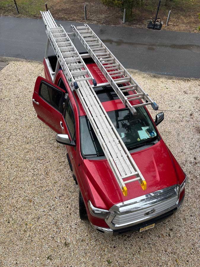 Red truck with two ladders on roof rack and driver's side door open, parked on gravel.