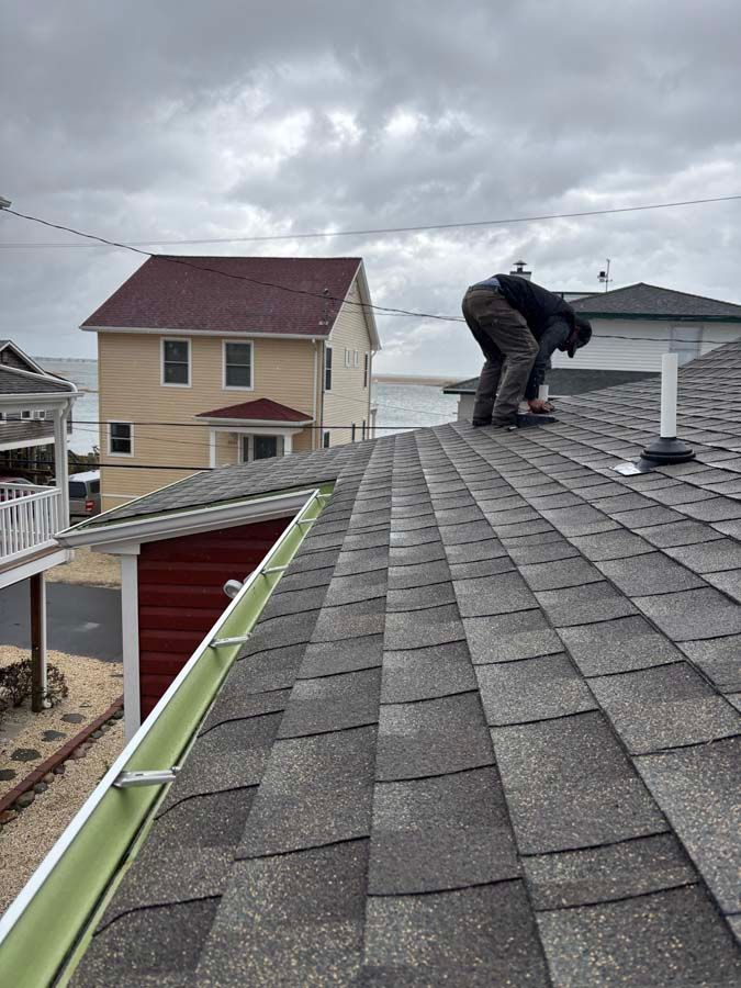Person on a shingled roof, working on it. Another house and water visible in the background. Cloudy sky.