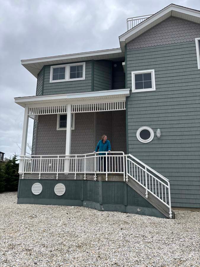 Person standing on the porch of a two-story blue-green house with white trim. Overcast day.