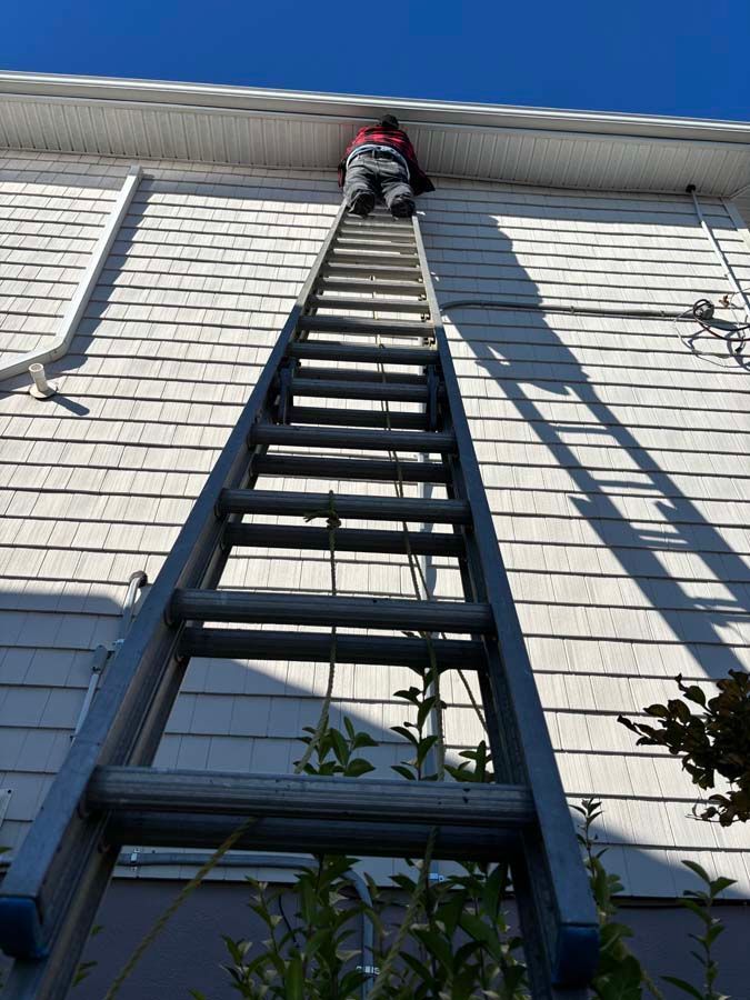 Person on ladder cleaning gutters on a white shingled house under a blue sky.