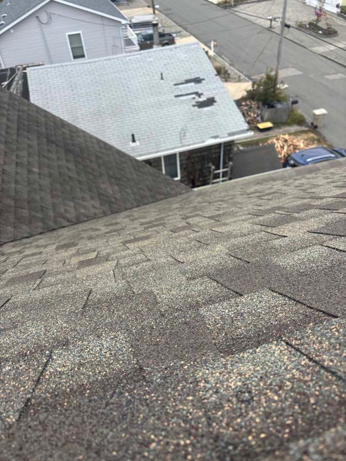 View of a weathered asphalt shingle roof, angled, with neighboring houses and street in the background.
