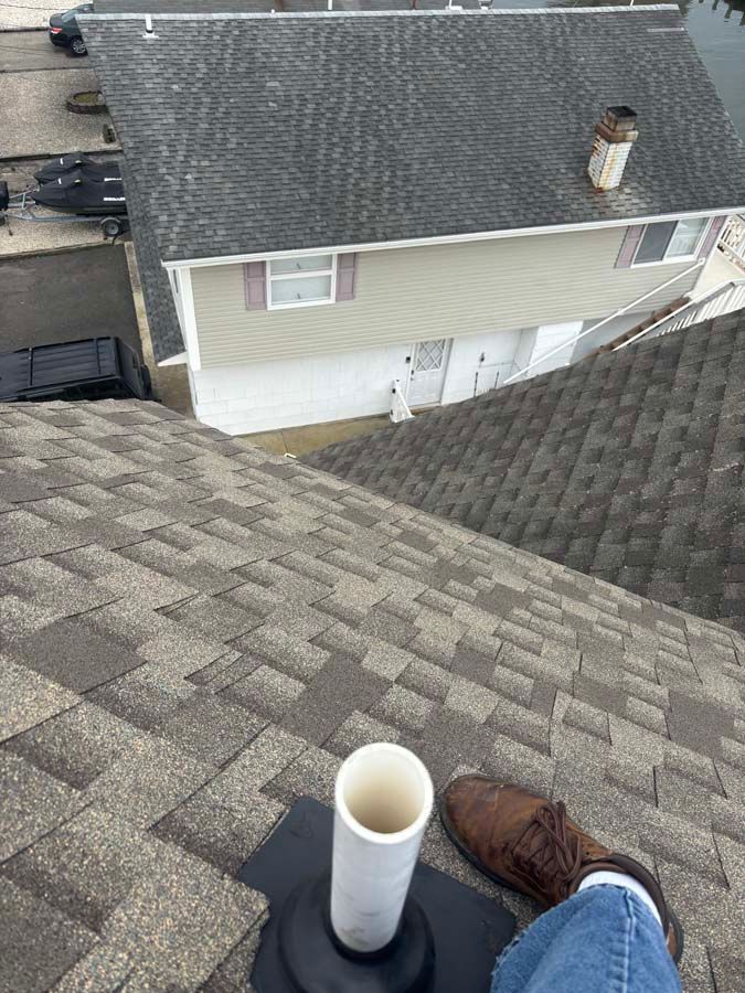 A person's foot on a shingled roof, next to a vent pipe. Building exterior visible in background.