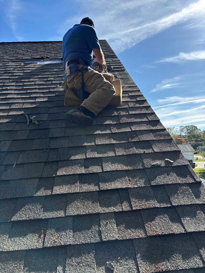 Person on a roof working with a box, blue shirt, brown pants. Sunny day, blue sky, house in the background.