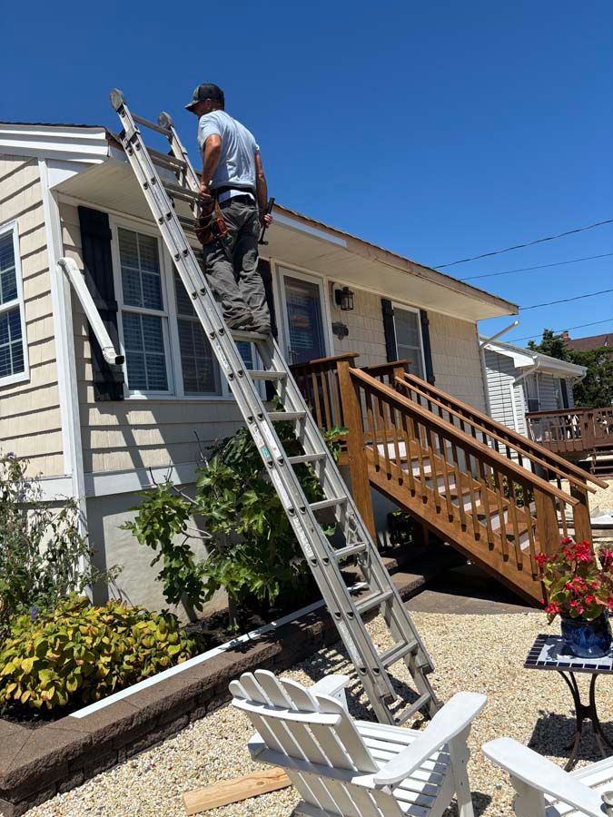 Person on a ladder, working on a house's roof. Sunny day, beige siding, white trim, wooden stairs.