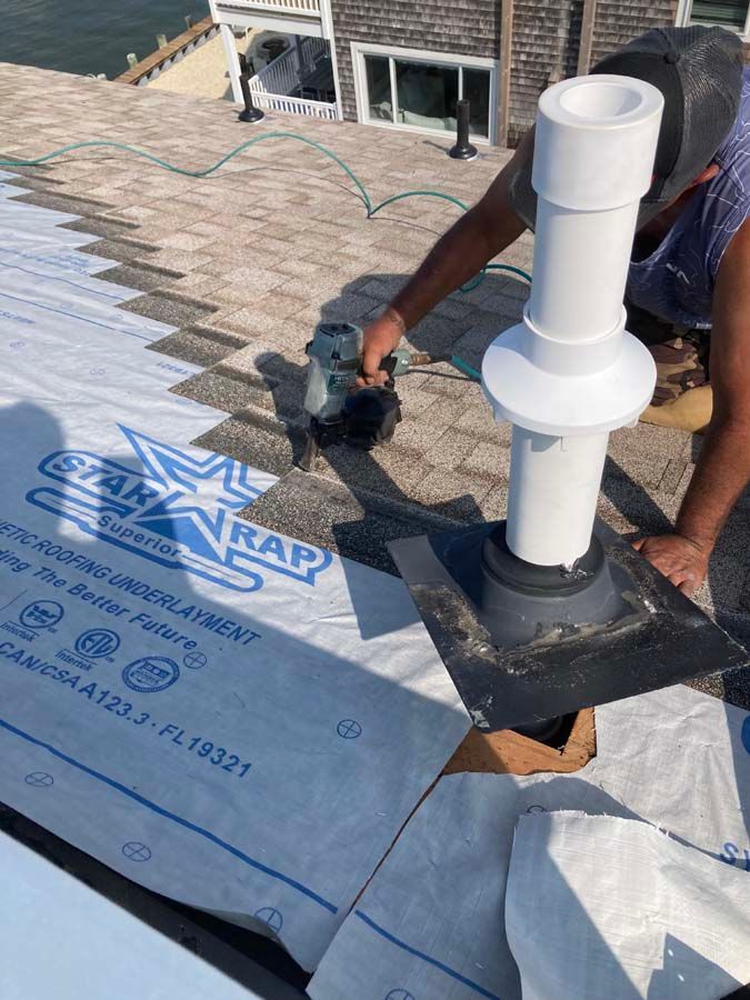 Roofer installing shingles around a vent pipe on a rooftop, using a nail gun.