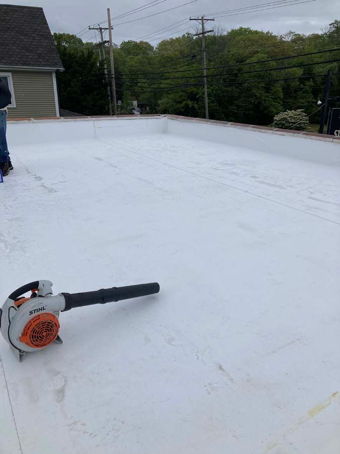 White roof being cleaned with a leaf blower. House and trees in background.