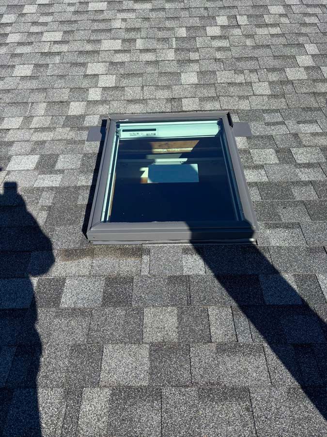 Skylight on a gray shingle roof, with a black frame and sunlight reflecting.