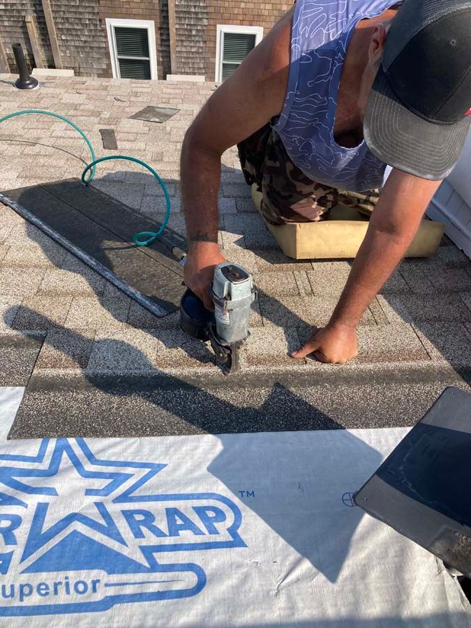 Roofer using a nail gun on a shingled roof, outdoors. The roofer wears a cap and blue patterned shirt.