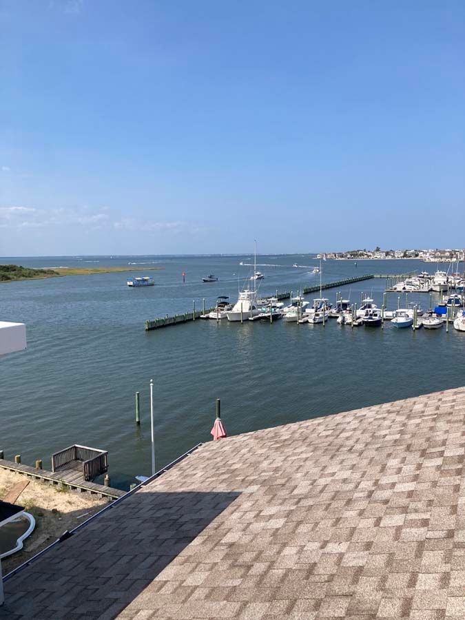 View of a marina with docked boats and blue water under a clear sky. A rooftop is in the foreground.