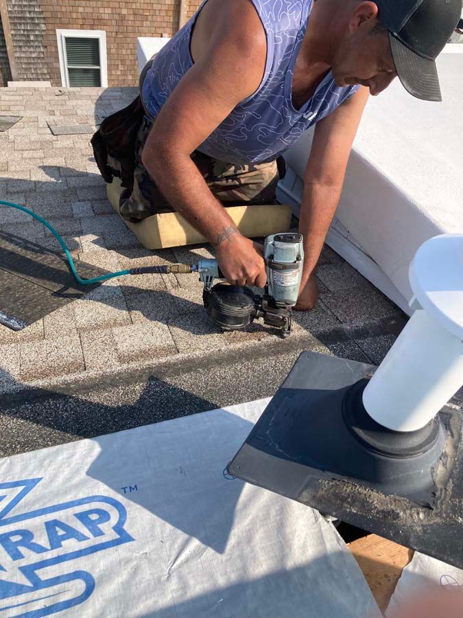 Roofer using a nail gun on a shingled roof, near a white pipe. A roll of building wrap sits nearby.