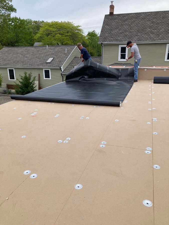 Two workers installing a black roofing membrane on a flat roof. Beige underlayment visible, residential setting.