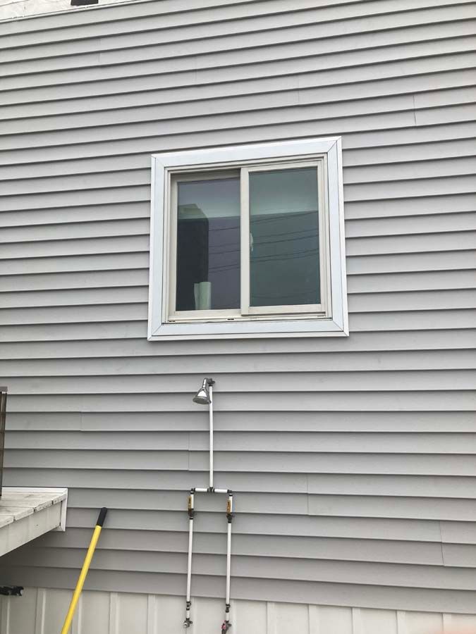 Gray siding with a white-framed window. Below, a shower head and two handles are mounted on the wall.