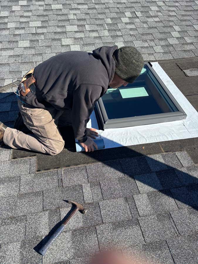 A person on a roof installing a skylight; holding tool, wearing work clothes, overcast.