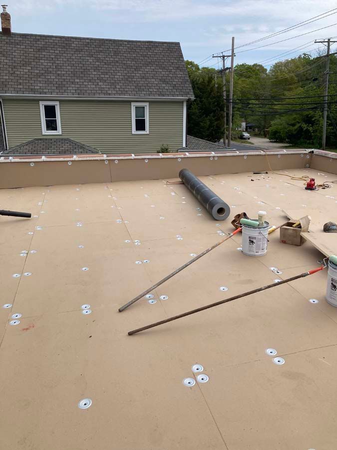 Flat roof with roofing materials and tools, a roll of roofing, and a house in the background.