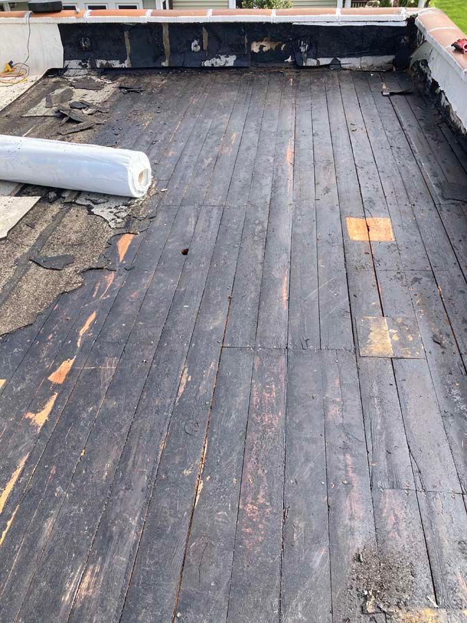 Overhead view of a flat roof with weathered wooden planks and sections of torn, black roofing material. A roll of white material is present.