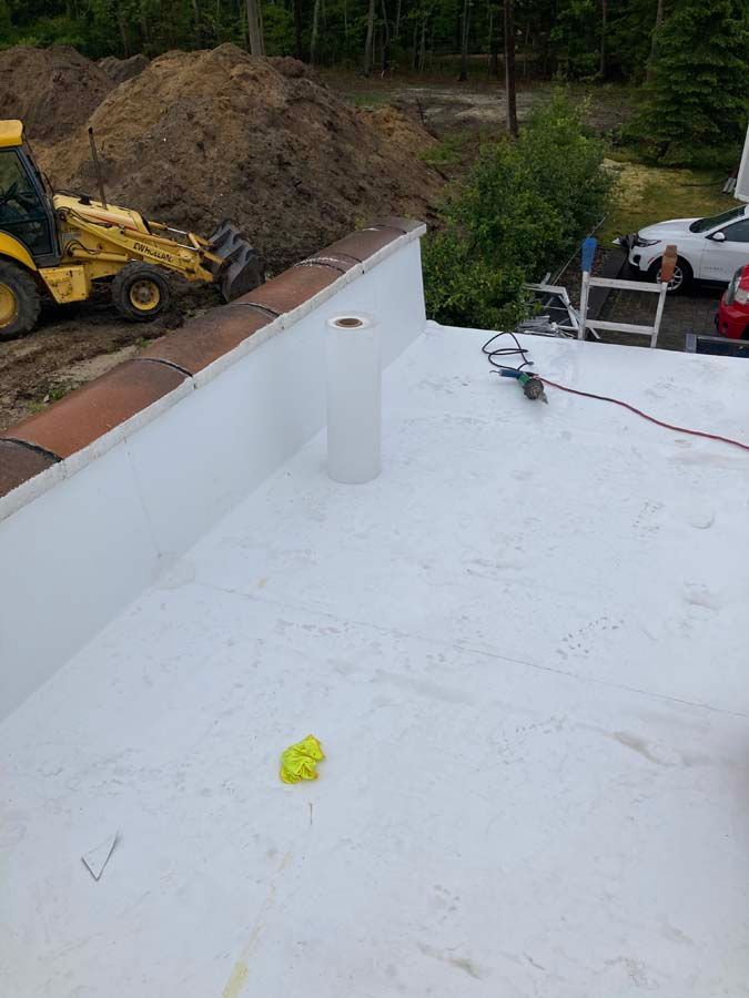 White flat roof with a vent pipe, next to a brick wall. A yellow backhoe is in the background.