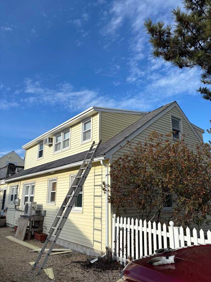 Yellow house with a ladder propped against the side, white picket fence, and blue sky with clouds.