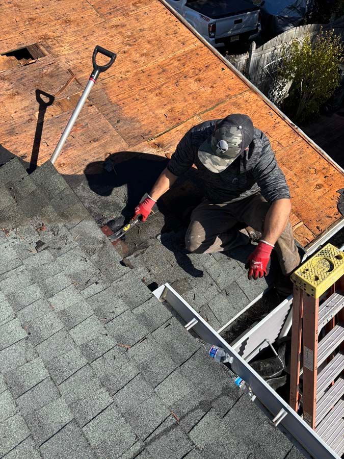 Roofer kneeling, hammering shingles on a roof. Ladder and tools visible.