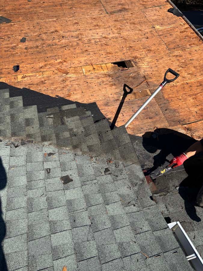 Roofer removing shingles.  Gray shingles in foreground, exposed wood roof in background. Tools on roof.