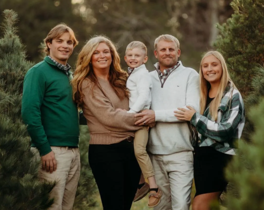 Smiling family of five standing outdoors in a wooded area, dressed in earthy tones.