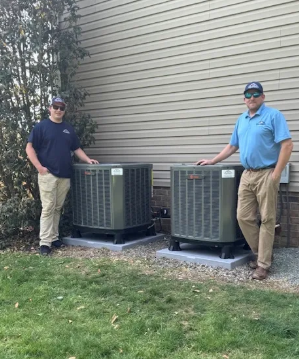 Two people stand beside outdoor HVAC units against a house wall.