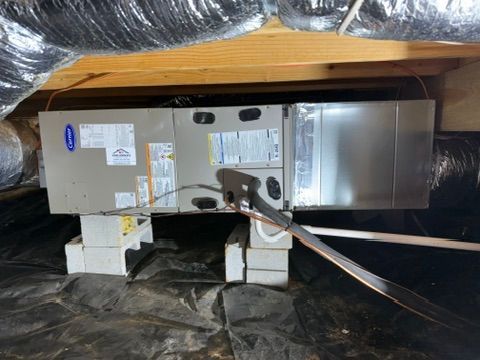 Large HVAC unit in a crawlspace, strapped to blocks under insulation and wooden joists.