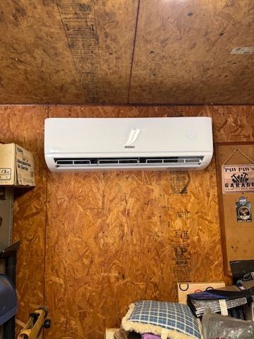 Wall-mounted white air conditioner on a plywood workshop wall with shelves and stored items nearby