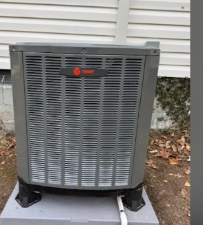 Gray Trane HVAC unit outside a house, sitting on black stands beside white siding.