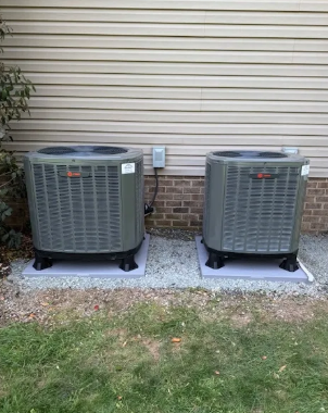 Two gray outdoor air conditioner units on concrete pads beside a beige house wall and brick foundation